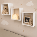 Three white square floating wall shelves mounted on a beige wall with cloud and star decals. The shelves display a teddy bear, a glowing cloud night light, and a framed ultrasound photo next to a plush bunny.