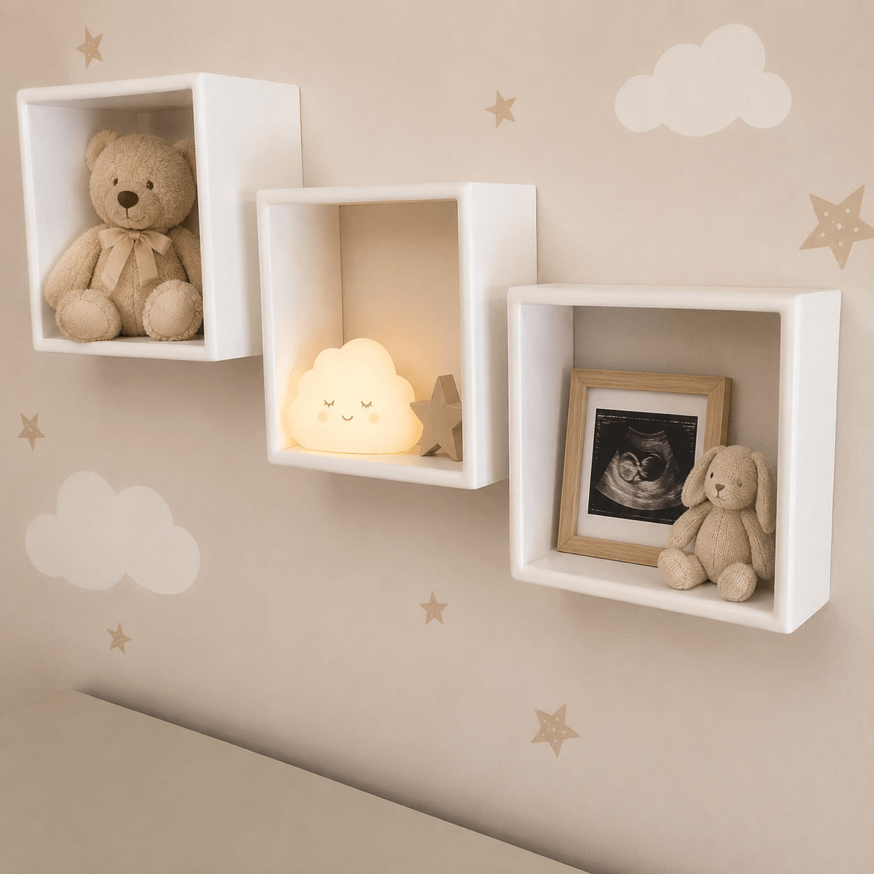 Three white square floating wall shelves mounted on a beige wall with cloud and star decals. The shelves display a teddy bear, a glowing cloud night light, and a framed ultrasound photo next to a plush bunny.
