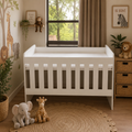 A white wooden toddler crib with vertical slats, styled in a warm, safari-themed nursery with giraffe and elephant wall decals, a window view, and plush toys on a woven rug.
