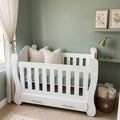 A white wooden baby crib with decorative pillows, set in a styled nursery with sage green walls, a window with sheer curtains, and a small desk area with a lamp.