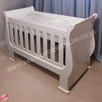 2in1 Sleigh wooden Cot White baby crib in a room with light blue curtains.