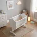 A white wooden baby crib with a curved headboard and footboard, styled in a soft, neutral nursery setting with a teddy bear inside, placed on a woven rug.
