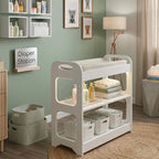 A white three-tier baby changing table with a mesh changing pad, styled in a nursery with sage green walls, wooden floors, and storage baskets.