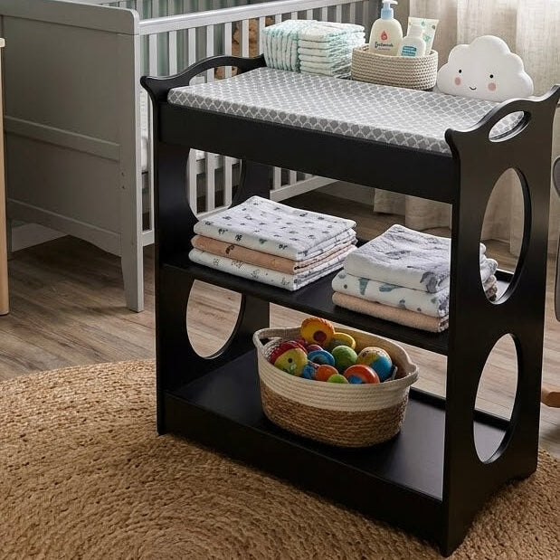A black wooden baby changing table with three tiers and decorative side cutouts, styled in a nursery with a gray crib, folded blankets, a basket of colorful balls, and a patterned changing pad on top.