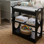 A black wooden baby changing table with three tiers and decorative side cutouts, styled in a nursery with a gray crib, folded blankets, a basket of colorful balls, and a patterned changing pad on top.