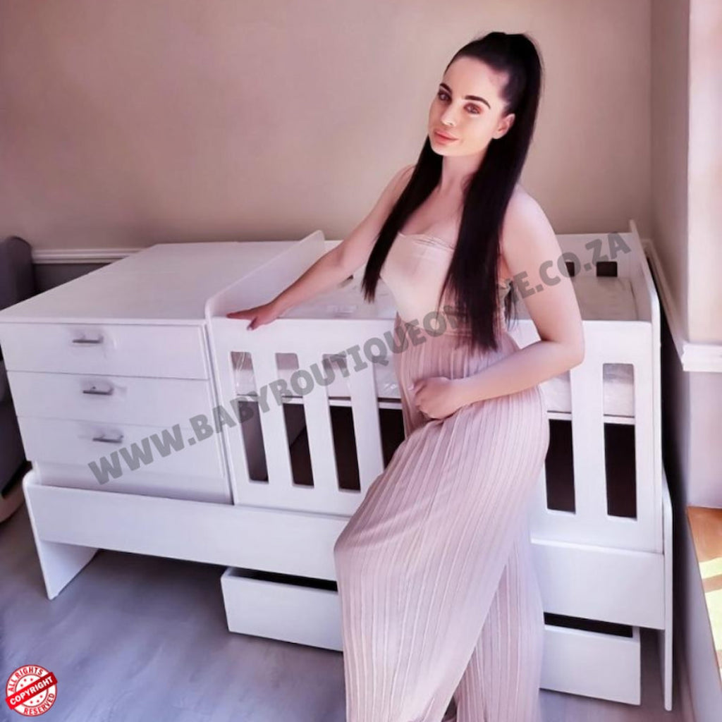 Woman in a light dress standing next to a Babybedroominabox5-in-1Cot white crib in a room with neutral walls.