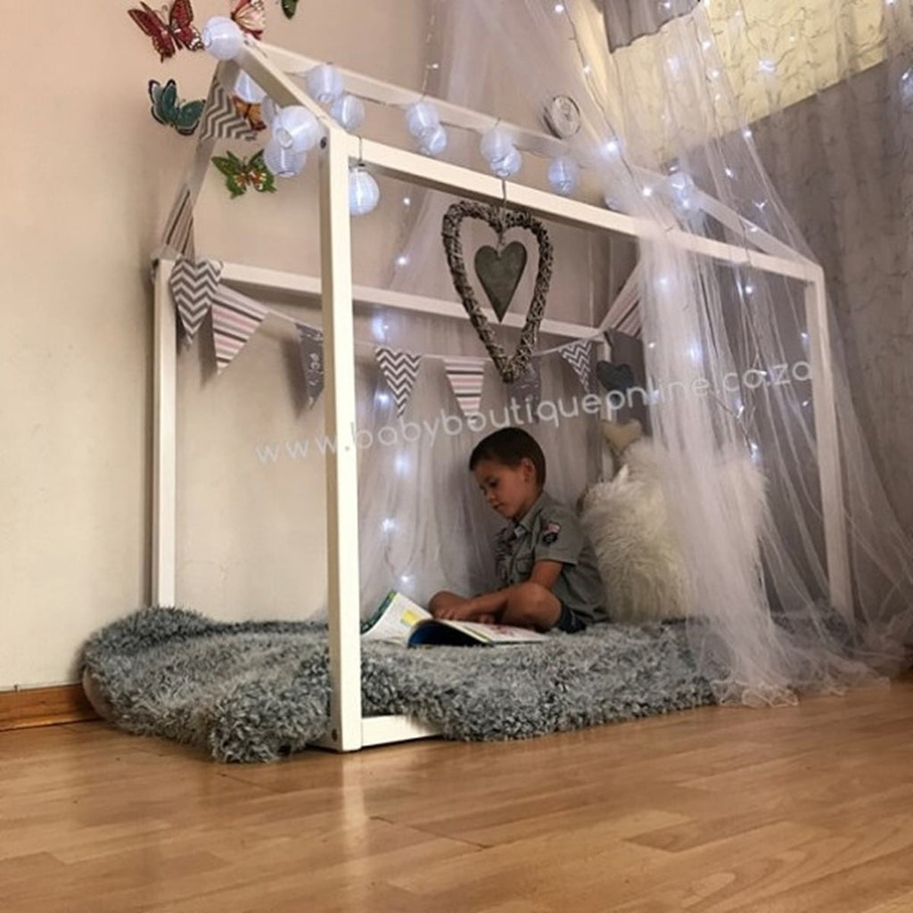 Child sitting on a DIY playhouse made of a crib frame with curtains and lights, reading a book.