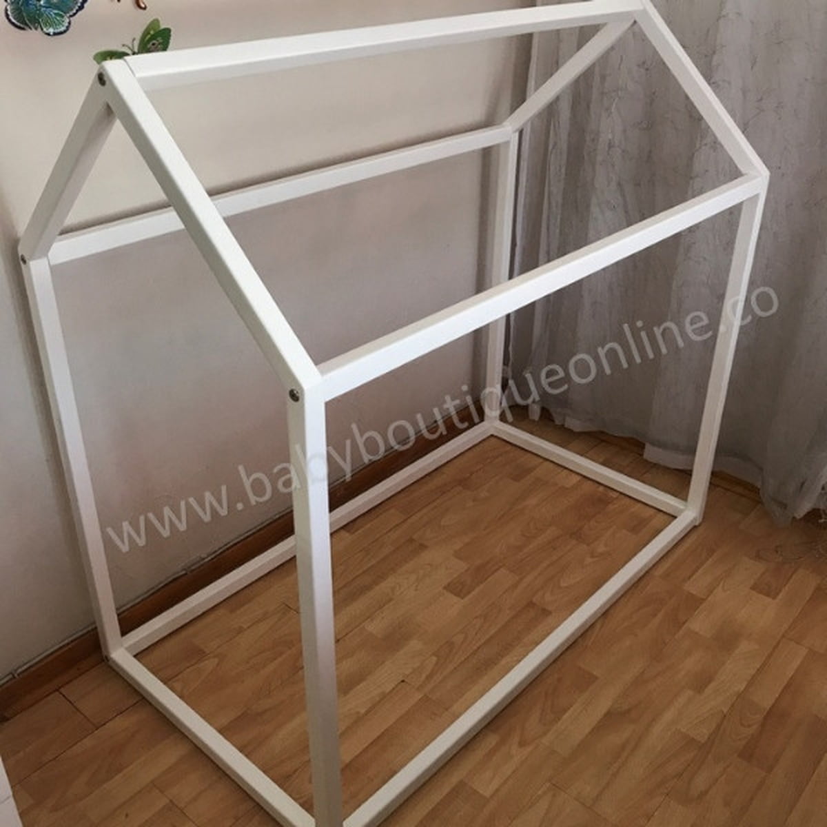 White metal frame house-shaped crib on a wooden floor with a neutral wall background.