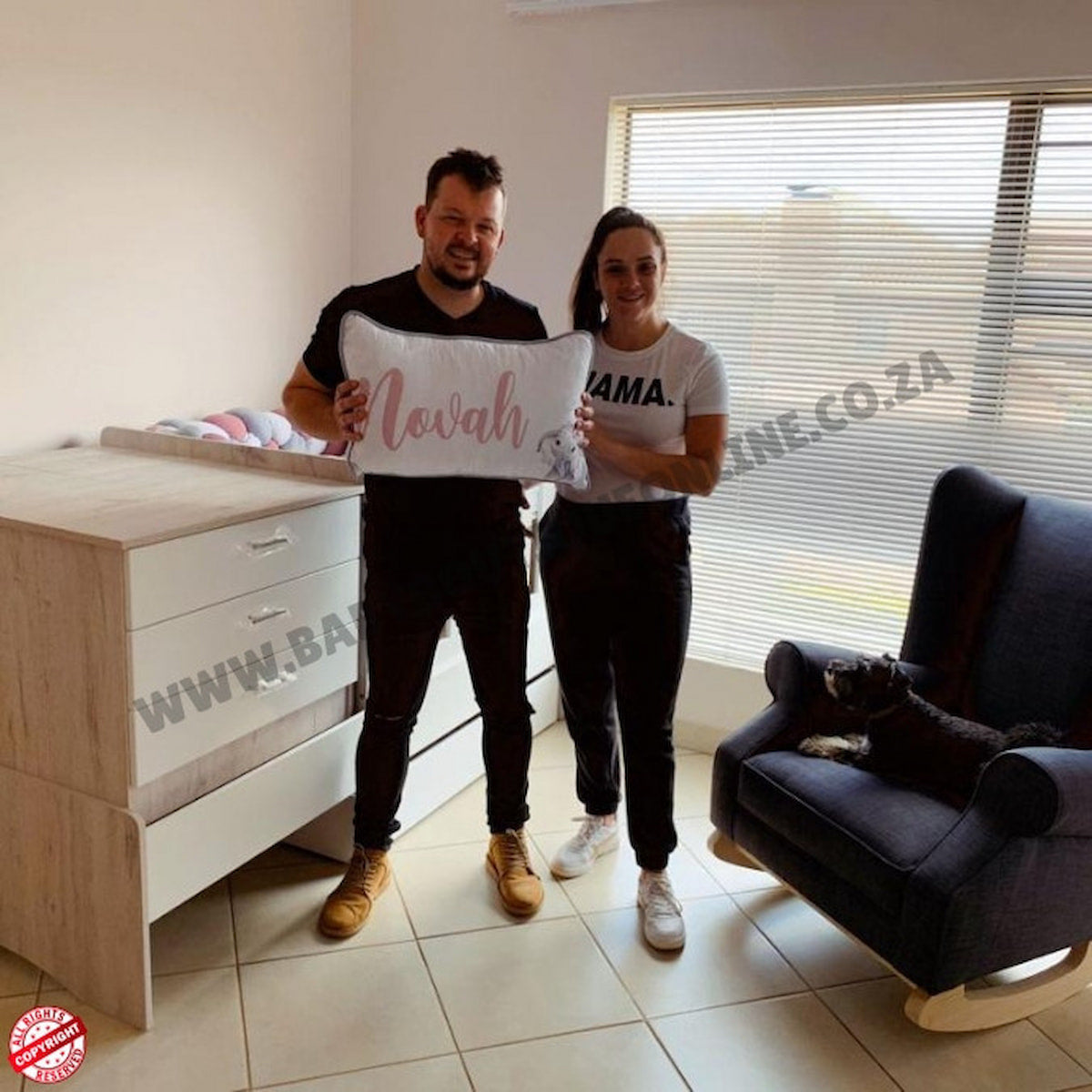 Juan Boucher and Wife holding a pillow with 'Novah' written on it in a room with furniture and a window.