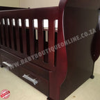 Sleigh Cot with Drawers Mahogany with drawers on a light wooden floor against a white wall.