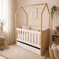 A house-shaped wooden toddler bed with a white slatted crib interior and base drawer, styled in a soft neutral nursery with plush toys, a rug, curtains, and framed bunny artwork.