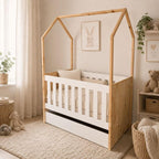A house-shaped wooden toddler bed with a white slatted crib interior and base drawer, styled in a soft neutral nursery with plush toys, a rug, curtains, and framed bunny artwork.