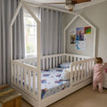 A white house-shaped canopy toddler bed with a slatted guard rail, shown in a child's bedroom with gray curtains, a ceiling fan, and a child in a pink onesie standing beside it.
