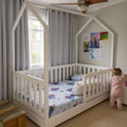 A white house-shaped canopy toddler bed with a slatted guard rail, shown in a child's bedroom with gray curtains, a ceiling fan, and a child in a pink onesie standing beside it.