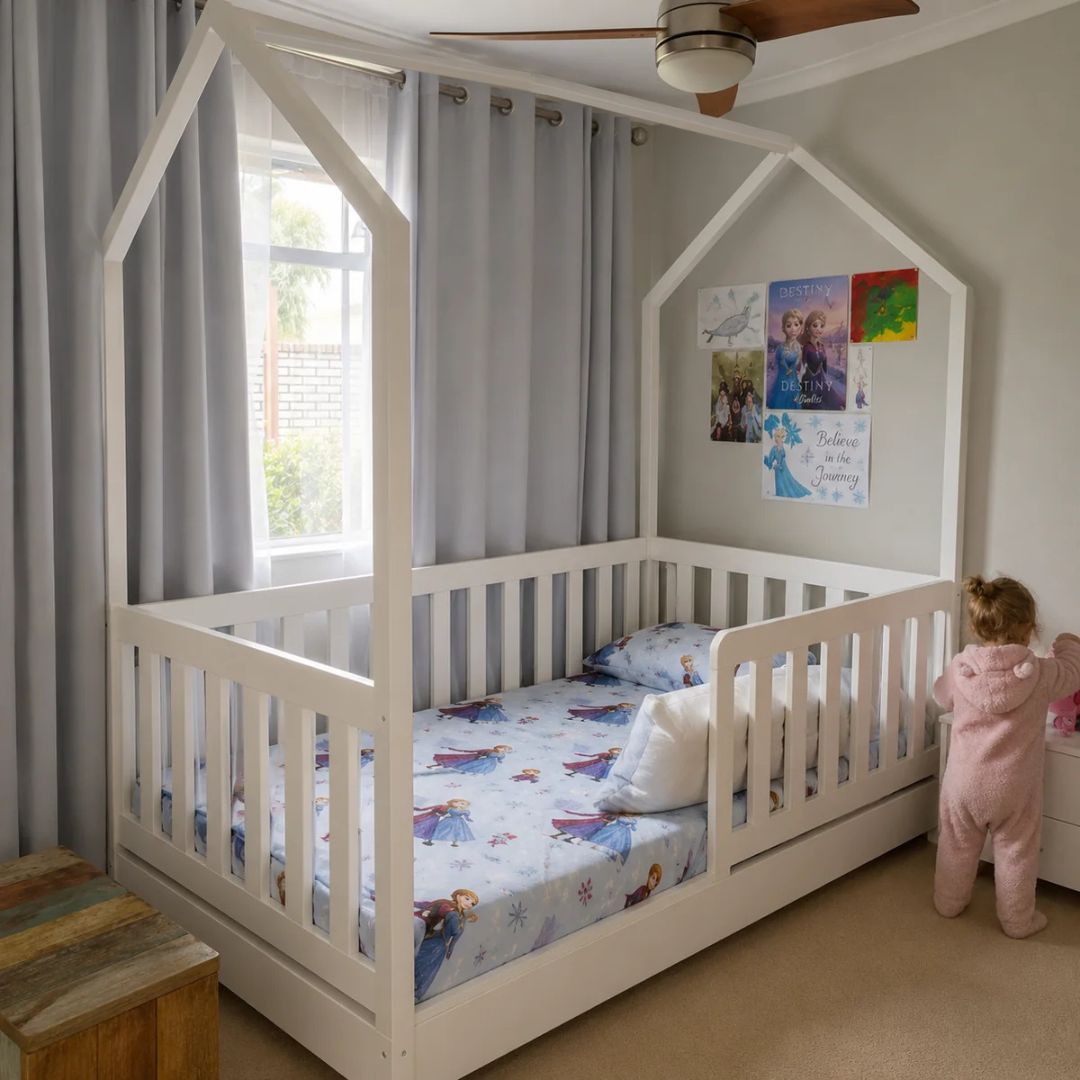 A white house-shaped canopy toddler bed with a slatted guard rail, shown in a child's bedroom with gray curtains, a ceiling fan, and a child in a pink onesie standing beside it.