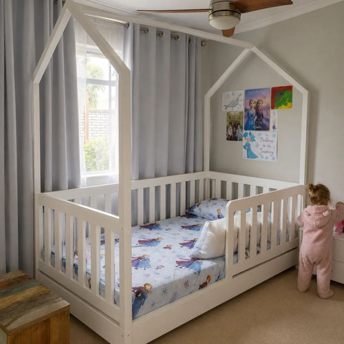 A white house-shaped canopy toddler bed with a slatted guard rail, shown in a child's bedroom with gray curtains, a ceiling fan, and a child in a pink onesie standing beside it.