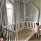 White children's playhouse with a small child peeking out from behind a curtain.