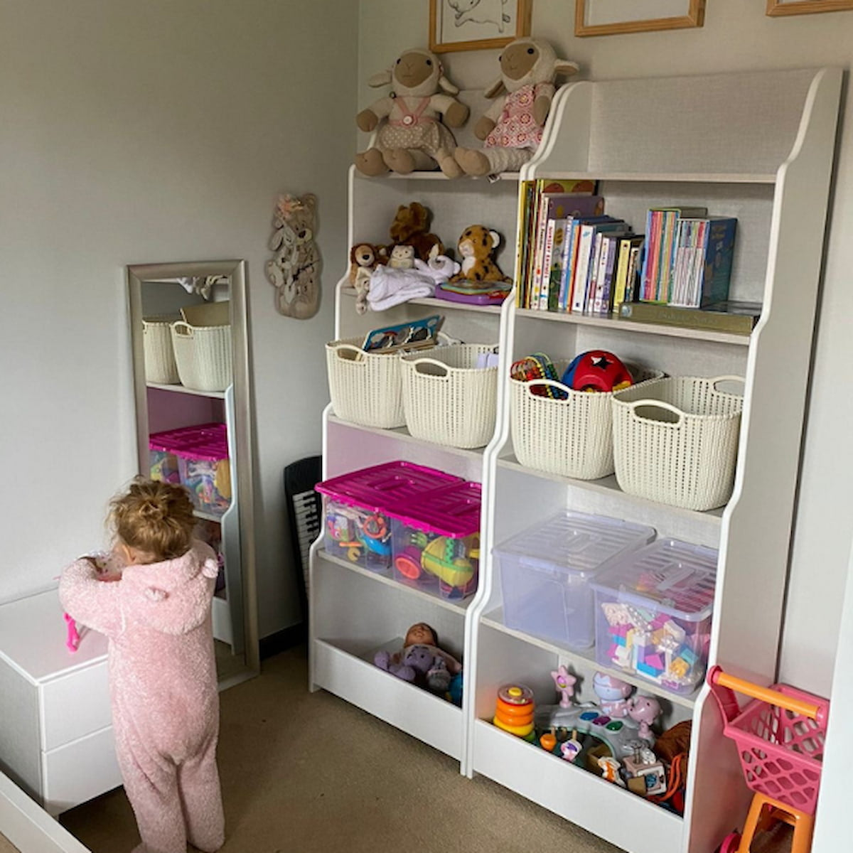 Child in a pink onesie standing next to a white shelving unit filled with toys and books in a room.