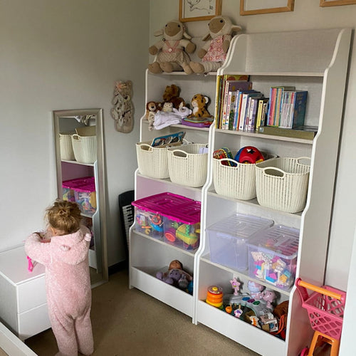 Child in a pink onesie standing next to a white shelving unit filled with toys and books in a room.