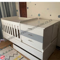 BabyBedroomInaBox5in1Cot White crib with changing table attached in a room with patterned wallpaper and curtains.