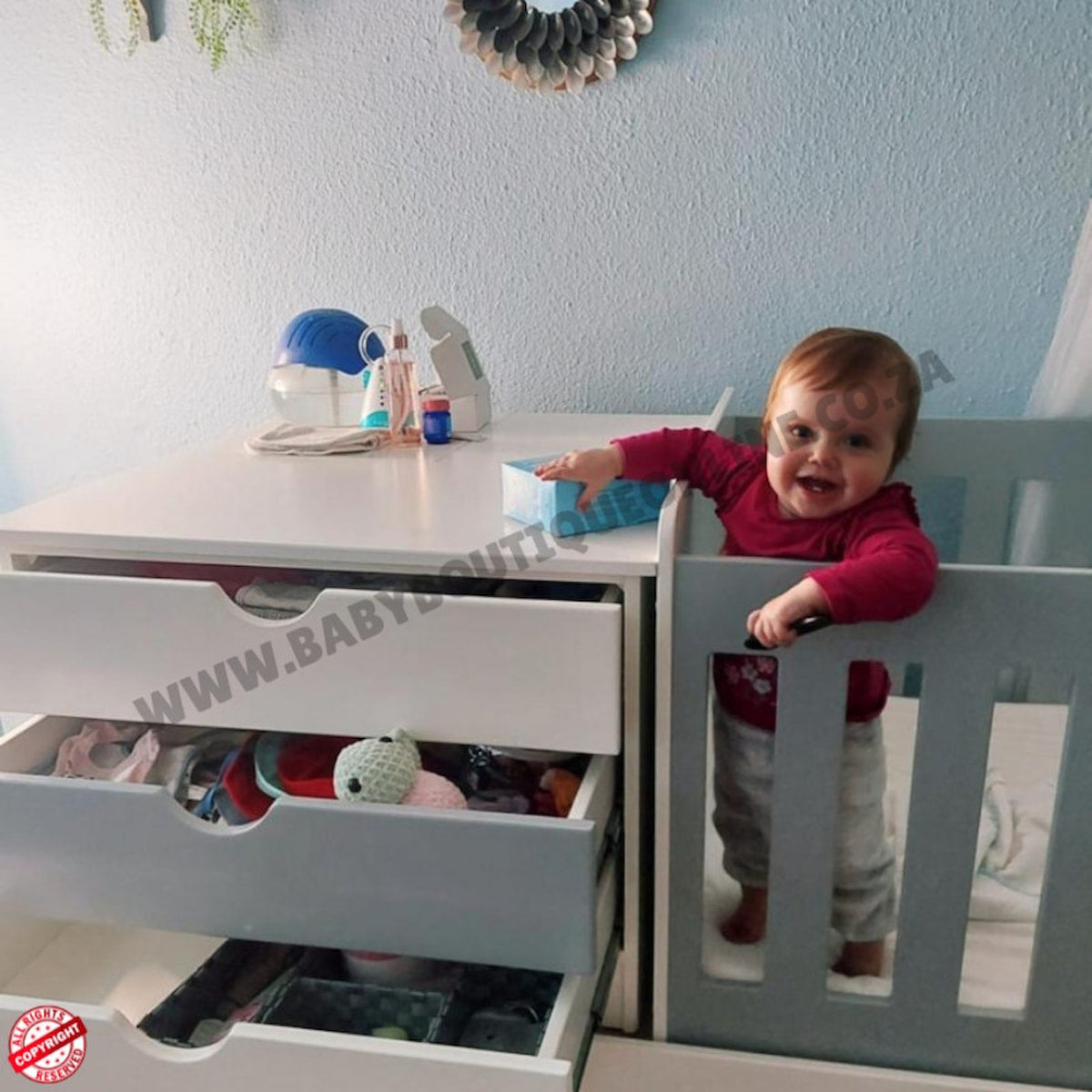Child playing with a toy in a BabyBedroominaBox5-in-1Cot crib next to a white dresser with open drawers.