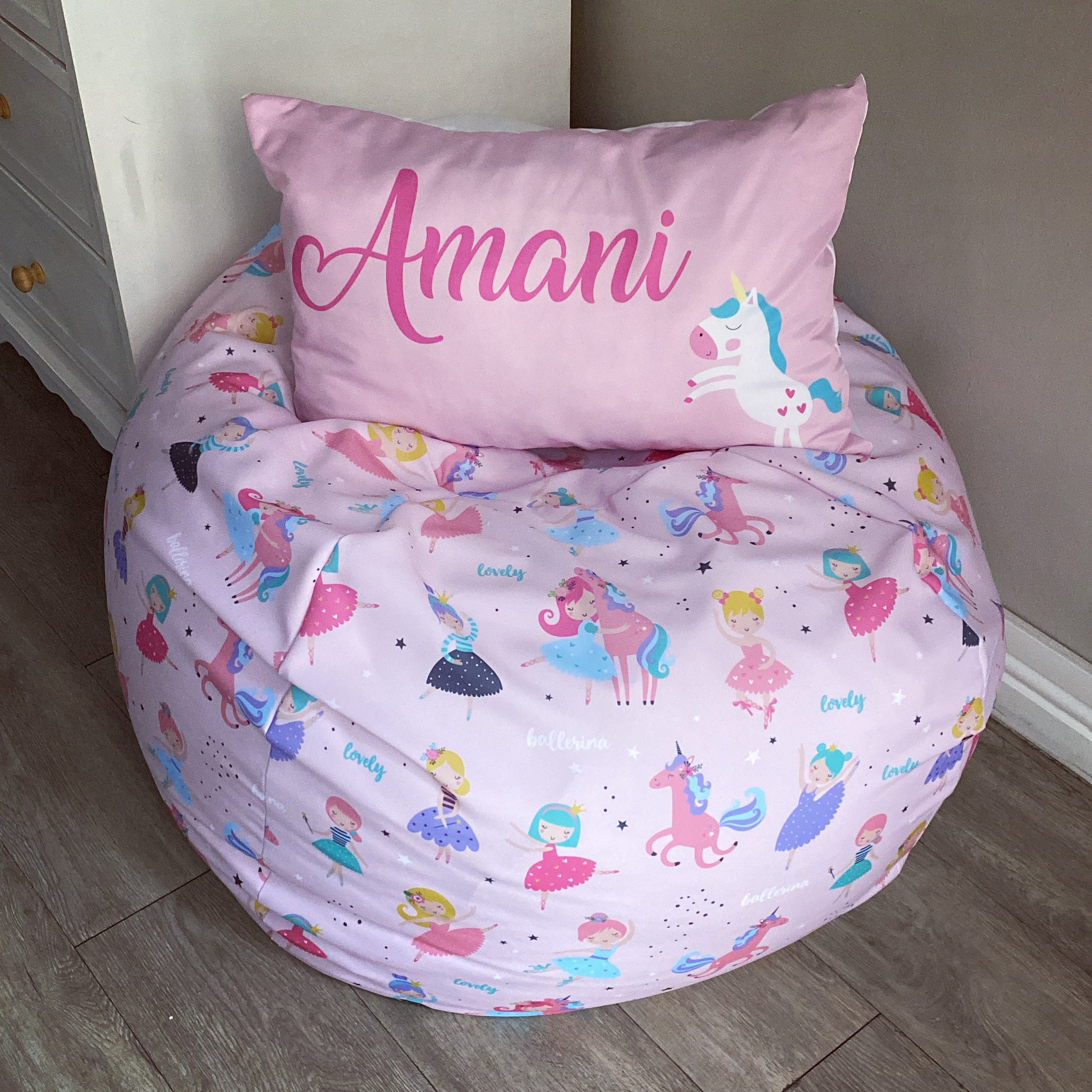 Children's bean bag chair with unicorn and fairy pattern and matching pillow with 'Amani' on a wooden floor.