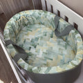 A round baby bassinet cushion set with a geometric patterned fabric in shades of sage green, yellow, and white, featuring gray straps, shown inside a white wooden bassinet.