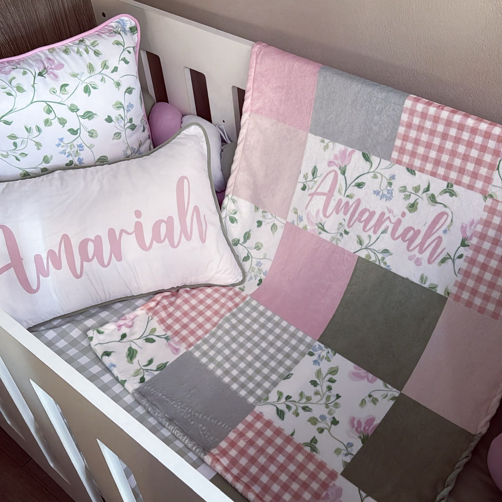 A pink and white patchwork baby bedding set in a white crib, featuring a quilt with floral and gingham patterns, a matching pillow, and a personalized name pillow that reads 'Amariah'.