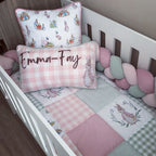 A baby bedding set styled in a white crib, featuring a pink and white gingham patchwork quilt, matching pillows with bunny illustrations and the personalized name 'Emma-Fay' in script, a green gingham pillow, and a pink and green bubble bumper.