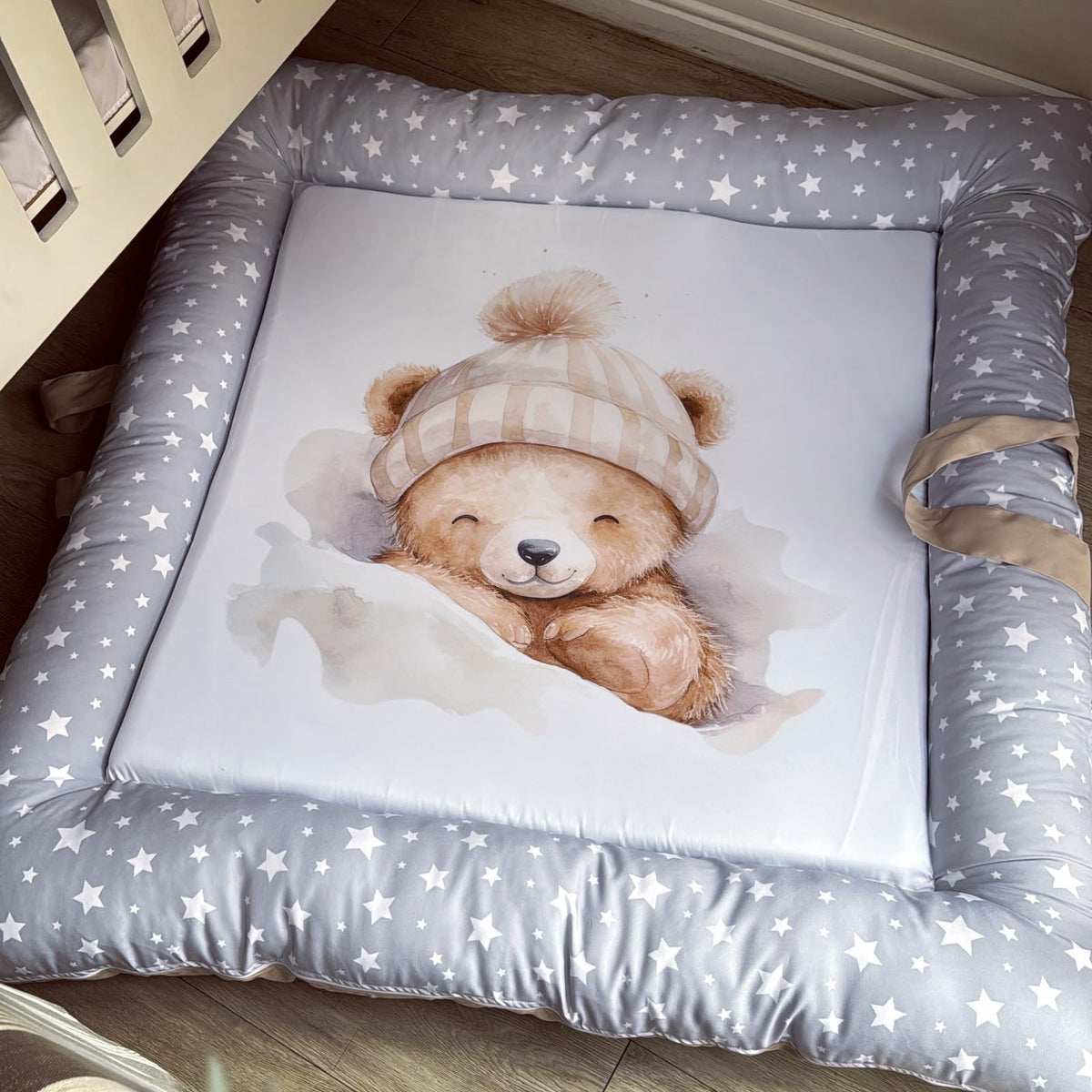 A padded baby floor mat or play mat with a gray star-patterned border and a white center, featuring a watercolor illustration of a teddy bear wearing a cream knit hat. The mat is placed on a wooden floor next to a crib.