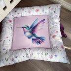 A square decorative cushion with a colorful hummingbird illustration on a light pink center panel, surrounded by a white floral border, photographed on a wooden floor.