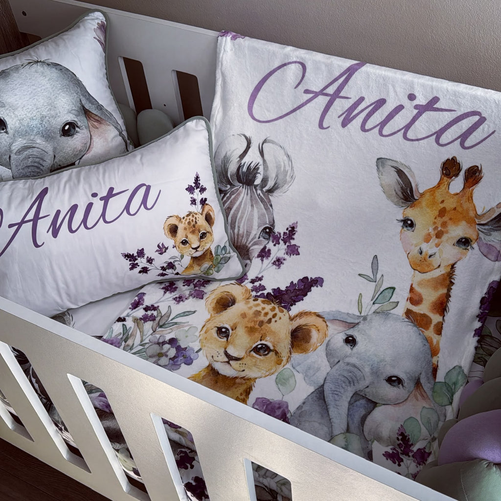 Custom safari-themed crib bedding set in a white wooden crib, featuring a duvet cover and matching pillowcases with watercolor illustrations of an elephant, giraffe, lion cub, and zebra, personalized with the name 'Anita' in purple script.