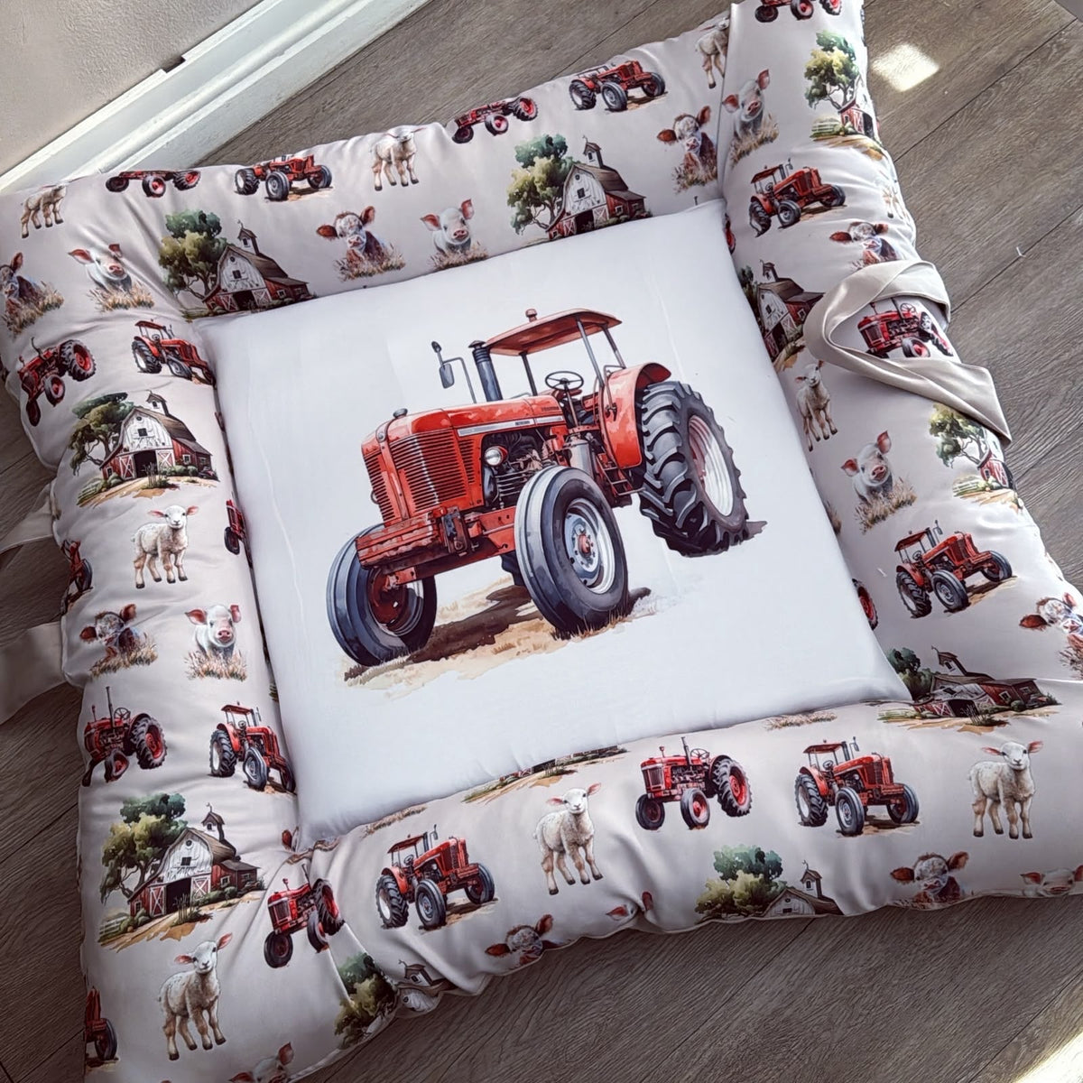 A square farm-themed pet bed with a white background and a printed border of red tractors, cows, sheep, barns, and trees, featuring a large central white square patch with a red tractor illustration.