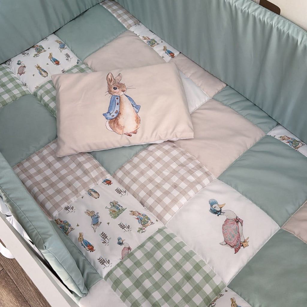 A baby bedding set in a crib, featuring a patchwork quilt in sage green, beige, and white with Peter Rabbit illustrations, and a matching pillow with a central Peter Rabbit print.