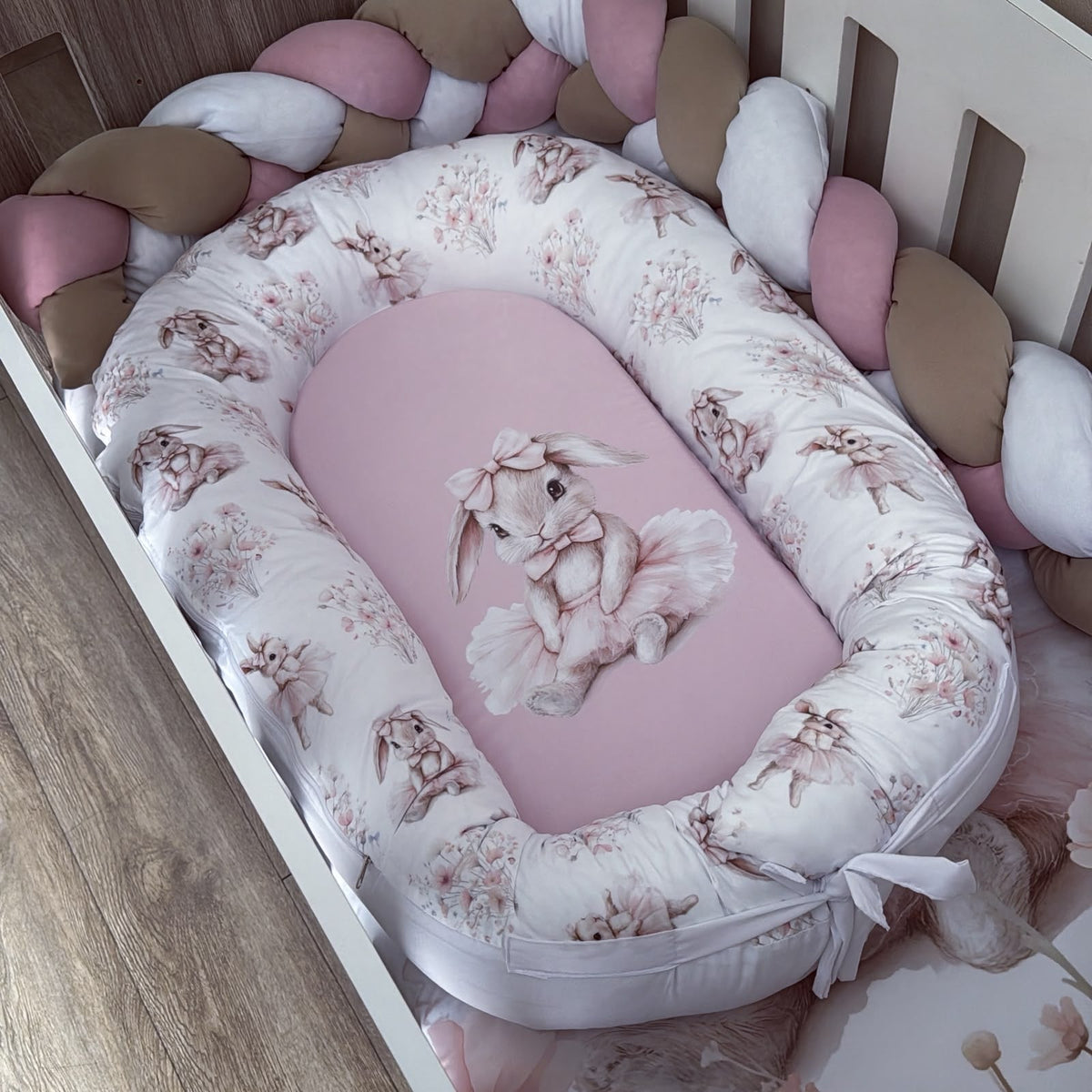 A baby bassinet bedding set featuring a pink bunny-themed mattress cover and a white bumper pad with a dusty pink bunny and floral print, styled inside a white wooden crib with braided pink and beige crib pillows.