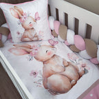 A white crib bedding set featuring a watercolor-style bunny illustration with pink flowers and a butterfly, including a fitted sheet, pillowcase, and chunky crib bumper in soft pink, beige, and white.