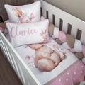 A white crib bedding set styled in a nursery, featuring a watercolor bunny design, a pink polka-dot sheet, a pink and white striped bumper, a chunky braided cord, and two matching pillows including one with the name 'Clarice' in pink script.