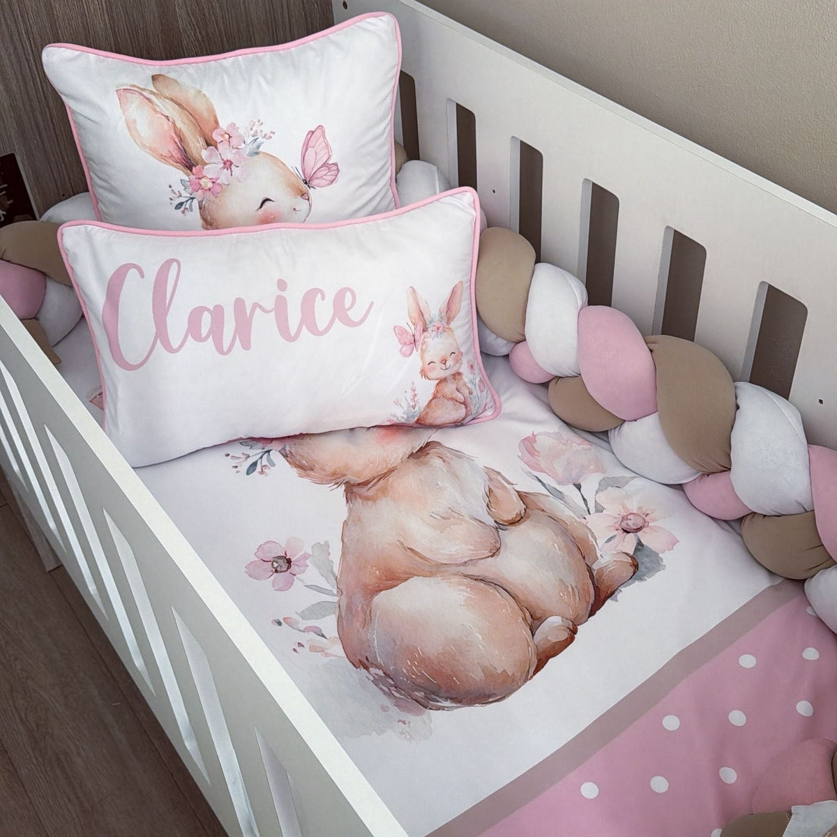 A white crib bedding set styled in a nursery, featuring a watercolor bunny design, a pink polka-dot sheet, a pink and white striped bumper, a chunky braided cord, and two matching pillows including one with the name 'Clarice' in pink script.