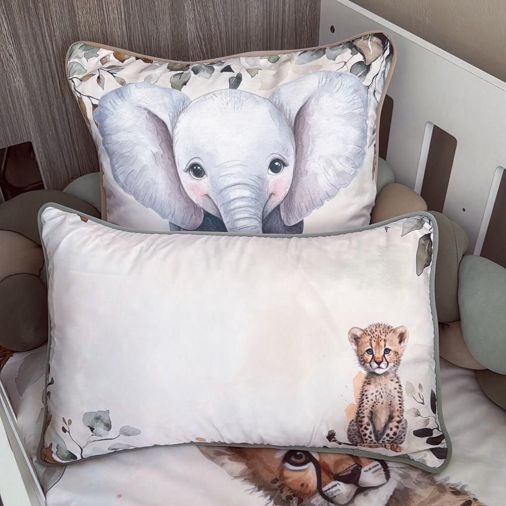 Two decorative pillows displayed on a bed in a nursery setting. The front pillow is off-white with a cute cheetah illustration and gray-green piping, while the back pillow features a large watercolor-style baby elephant with a patterned border.