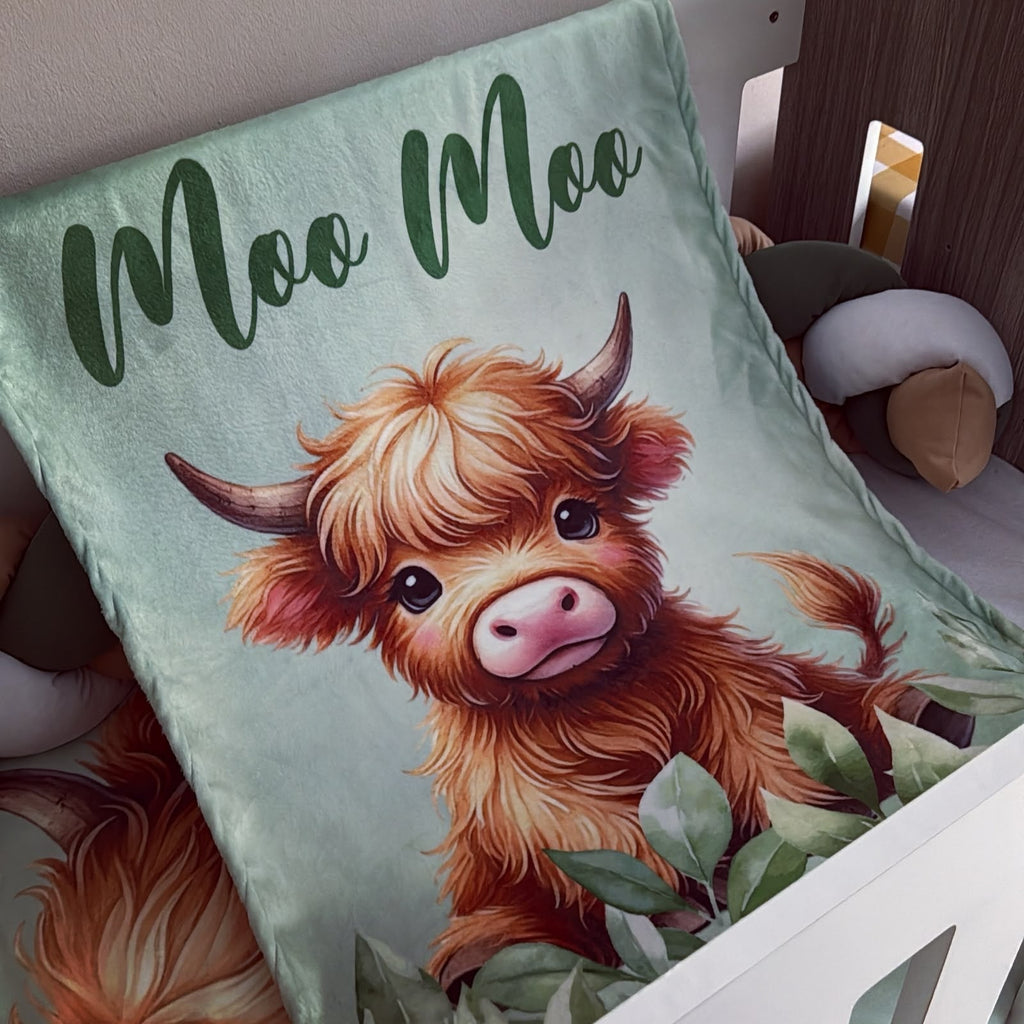 A soft fleece blanket draped over a white crib, featuring a cute illustration of a fluffy Highland cow with the text 'Moo Moo' in dark green script at the top.