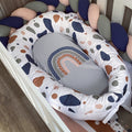 A baby bassinet cushion set in a white crib, featuring a padded oval bassinet cover with a white background and multicolor abstract shapes in navy, brown, and gray, surrounding a gray baby pillow with a rainbow design.
