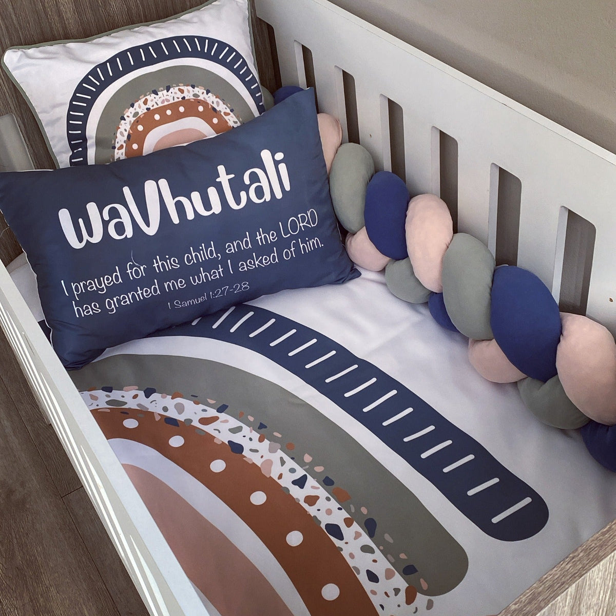 A styled nursery crib setup featuring a personalized navy blue pillow with white text reading 'waVutali I prayed for this child, and the LORD has granted me what I asked of him', a matching printed bumper cushion, and a chunky braided crib bumper in navy, blush pink, and gray.