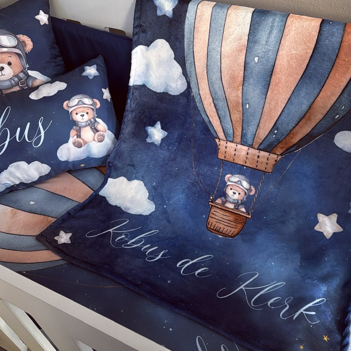 A cozy children's bedding set featuring a dark blue space-themed blanket and matching pillows with a watercolor hot air balloon, teddy bear, clouds, stars, and the personalized name 'Købus de Klerk' in white script.