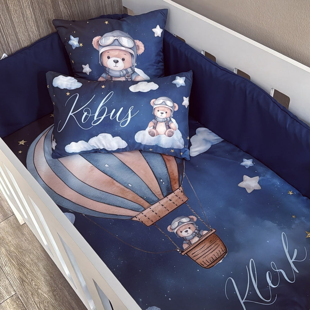 Custom baby bedding set in a crib, featuring a dark blue space-themed print with a teddy bear pilot in a hot air balloon, clouds, stars, and personalized names 'Kobus' and 'Kler' in white script.