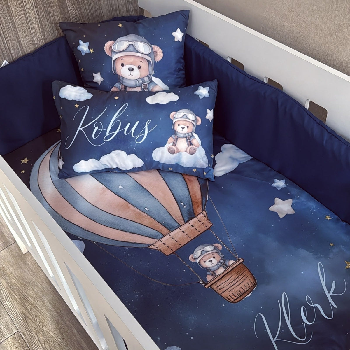 Custom baby bedding set in a crib, featuring a dark blue space-themed print with a teddy bear pilot in a hot air balloon, clouds, stars, and personalized names 'Kobus' and 'Kler' in white script.