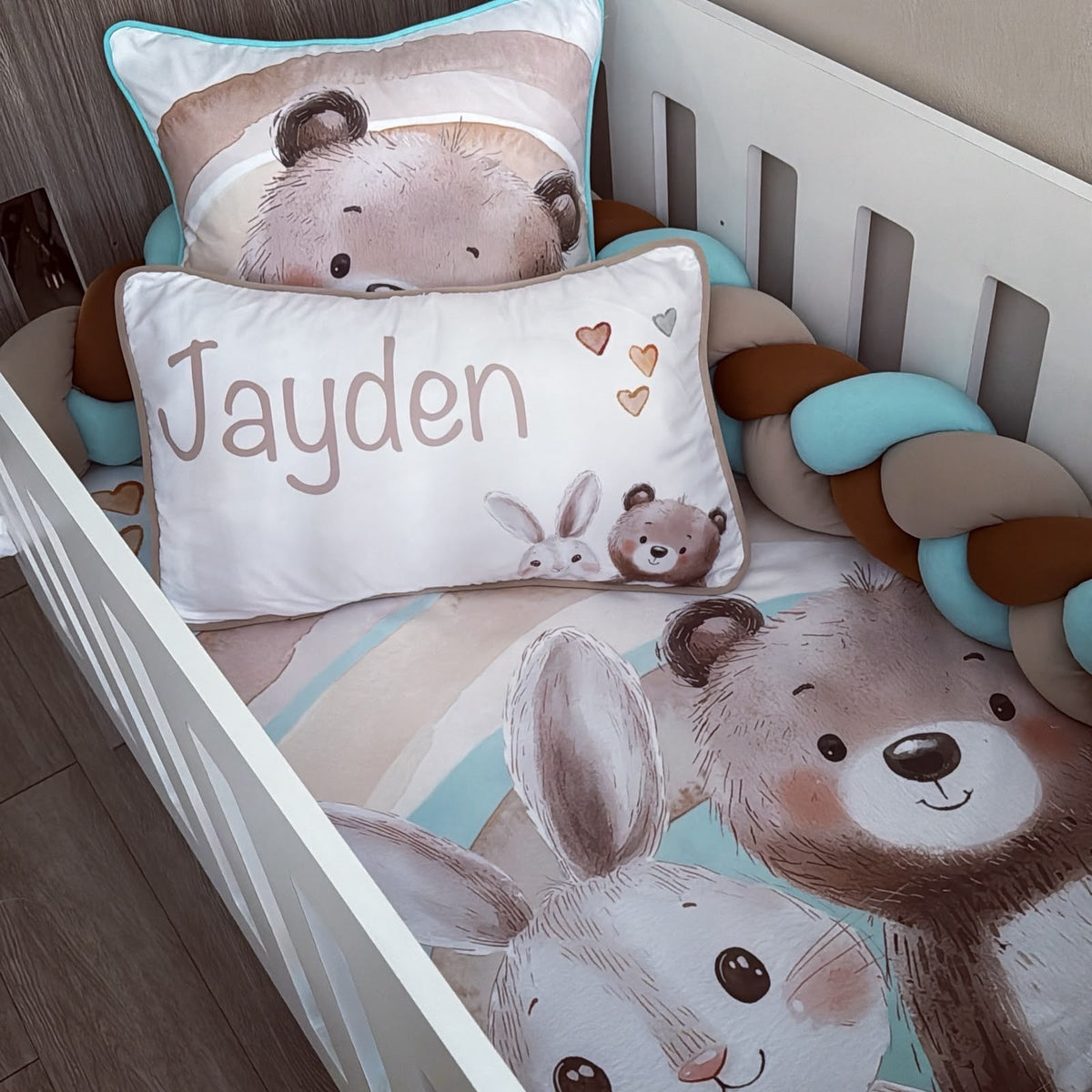 A personalized baby bedding set in a white crib, featuring a printed comforter and pillowcase with cartoon bear and bunny illustrations, a custom name pillow reading 'Jayden', and a chunky braided bumper in brown, beige, and light blue.