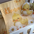 Baby blanket with duck design and 'Khensan' text, accompanied by a matching plush toy, on a crib.