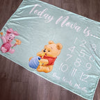 Baby milestone blanket with cartoon characters and numerical text on a wooden floor.
