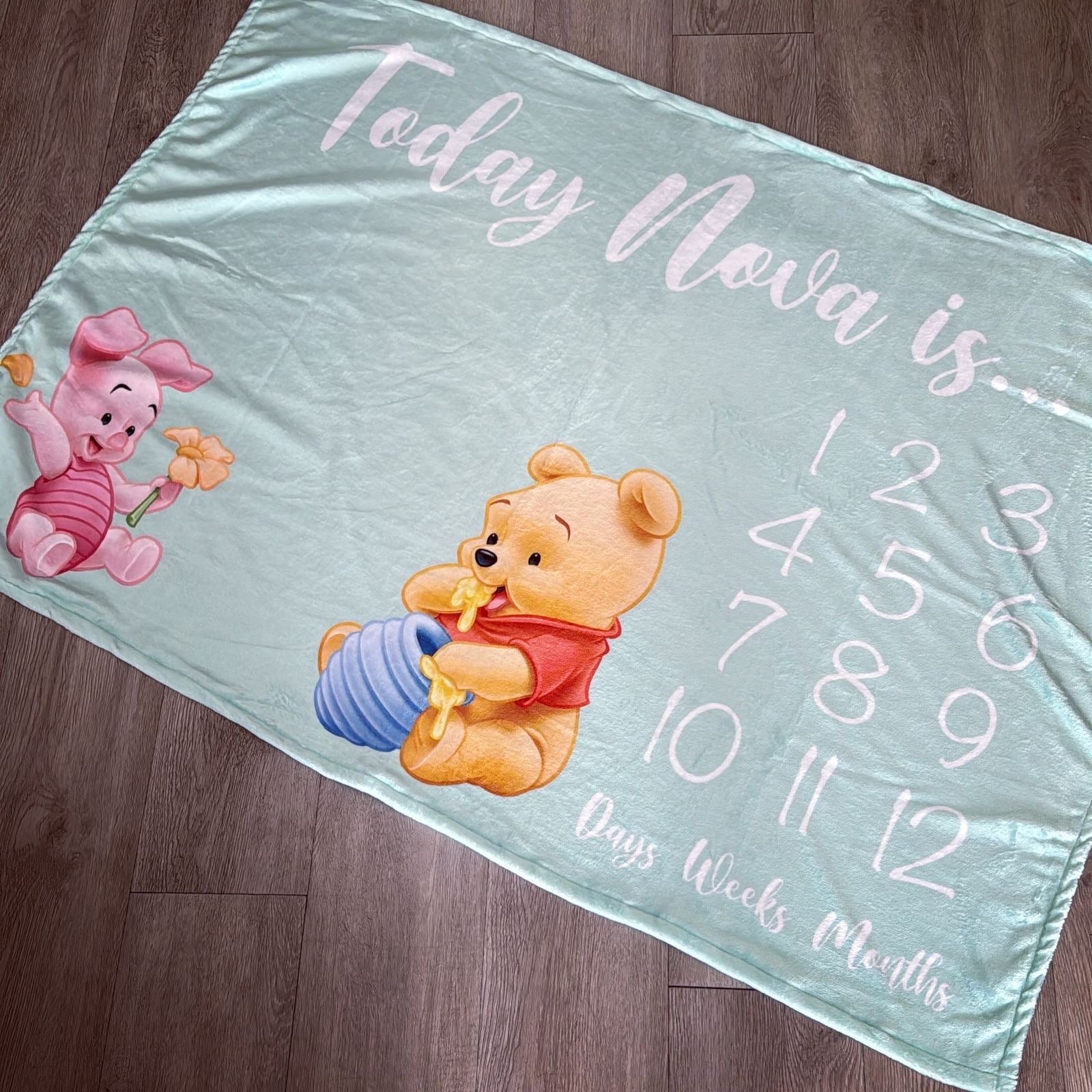 Baby milestone blanket with cartoon characters and numerical text on a wooden floor.