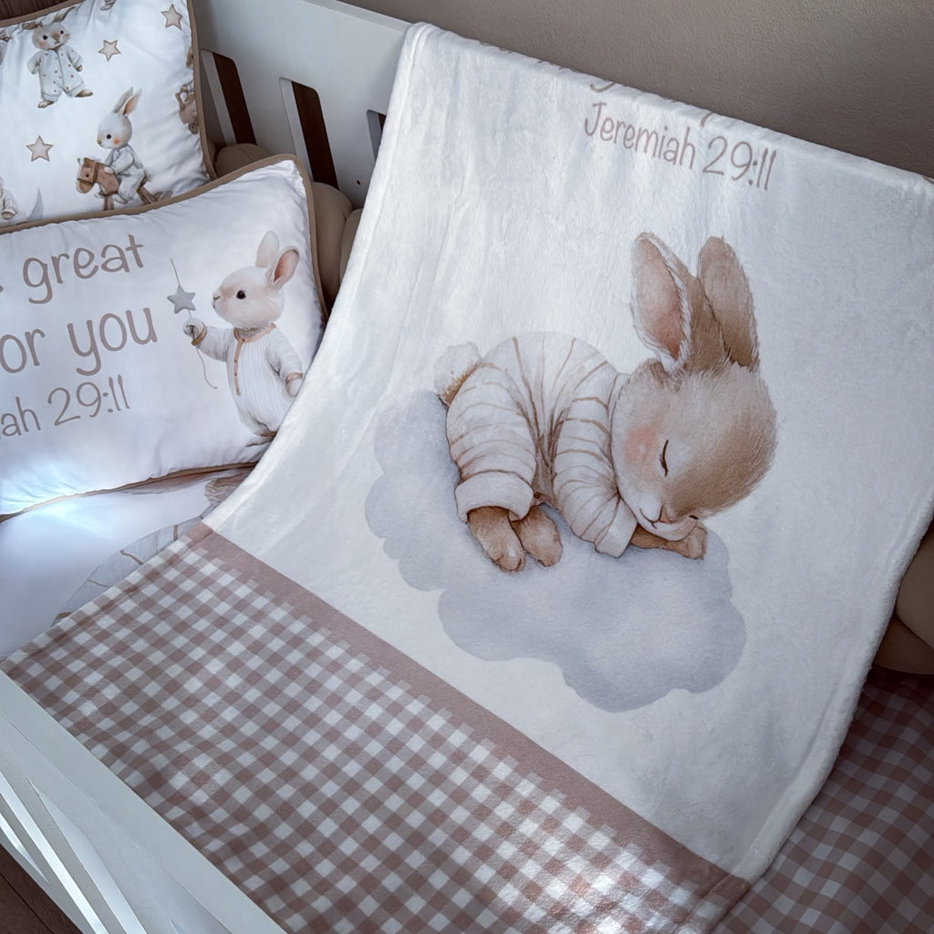 A baby bedding set featuring a white blanket with a sleeping bunny illustration and the text 'Jeremiah 29:11', a matching pillow with bunny graphics and text, and a pink-and-white gingham check blanket draped over a crib.
